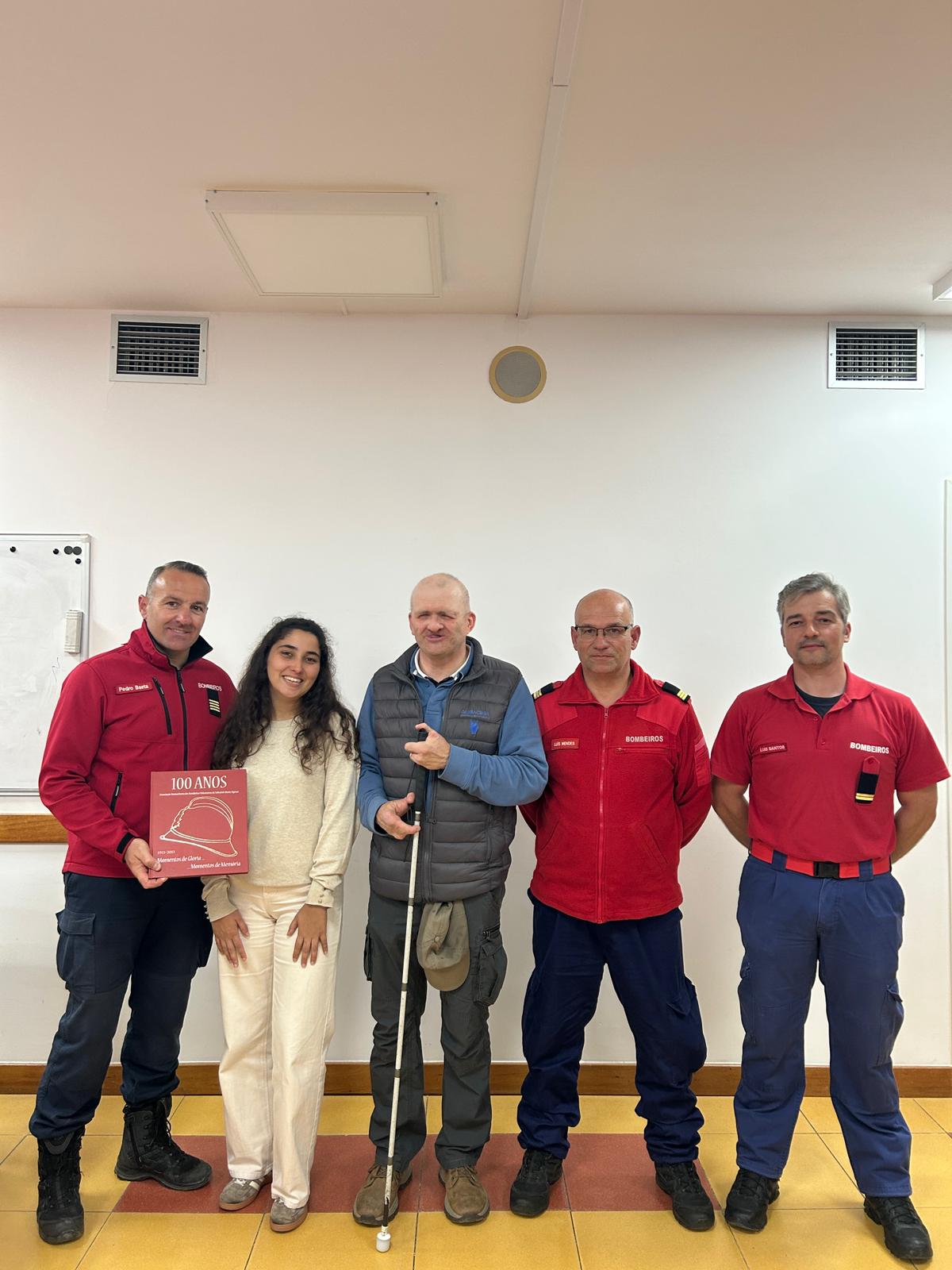Grupo de cinco pessoas em pé, alinhadas, contra uma parede branca, numa sala interior com chão de azulejos amarelos e uma faixa avermelhada.   Da esquerda para a direita: Um homem de pele clara, cabelo curto, com uniforme vermelho com a palavra “BOMBEIROS” e calças azul-escuras, a segurar uma placa vermelha. Uma mulher de pele clara, cabelo comprido e escuro, com roupa clara (camisola e calças), a sorrir. Um homem de pele clara, careca, com colete cinzento por cima de camisola azul e calças escuras, a segurar uma bengala branca. Um homem de pele clara, careca, com óculos, com uniforme vermelho “BOMBEIROS” e calças azul-escuras. Um homem de pele clara, cabelo curto grisalho, com polo vermelho “BOMBEIROS” e calças azul-escuras. Texto legível na placa vermelha: “100 ANOS”. Também há um desenho de um capacete e linhas menores de texto por baixo, mas não ficam totalmente legíveis nesta imagem.